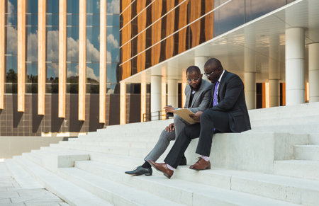 Business meeting outdoors. Two dark-skinned men in suits sit on the steps of a city building with a notebook and a laptop and have a conversation. Friendly support and brainstorming. Partnershipsの写真素材