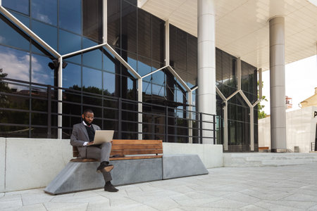 A dark-skinned man in a business suit is using a laptop outdoors. Working break. Online learning and education. Distance courses and communication.の写真素材