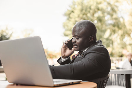 Portrait of black african american businessman in suits sitting at a table in a summer cafe with a laptop and talking on the phone outdoorsの写真素材