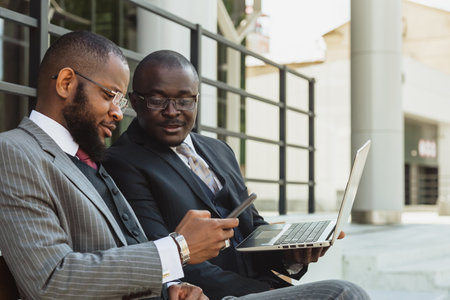 Business meeting of friends outdoors. Two dark-skinned men in suits are sitting on a bench near a city building with a laptop and talking. Partnership and corporate relationsの写真素材