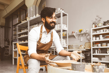 A young male potter is engaged in craft in his workshop on a potters wheel and makes a clay productの写真素材