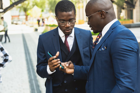 A couple of black African American businessmen friends in suits are talking while standing on the street. Outdoor business meetingの写真素材