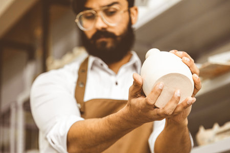 A young attractive male potter with a beard and mustache works in his workshop. Holds and inspects the finished productの写真素材