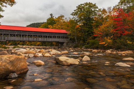 The red roof of the covered bridge at Albany, New Hampshire stands out against a cloudy sky and...