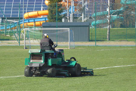 Worker on a large green lawn mower mows the grass on the football field. Landscape design and...