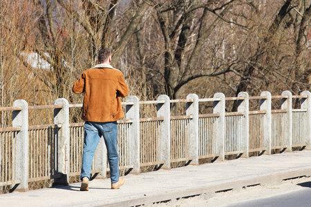 A young tall guy in jeans and leather sheepskin coat walks along the sidewalk with a confident fence. Confident man implements the plans. Hurry to a meeting. Modern urban demi-season clothing.の写真素材