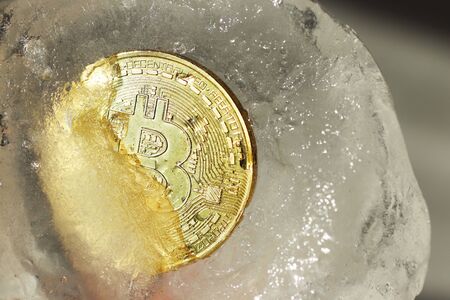 Bitcoin gold coin frozen in half in a piece of ice on a gray background. The freezing of financial...