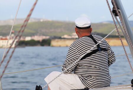 Yachtsman in marine clothing looking into the distance sitting on board a sailing yacht. Sea fishing from the ship. Peaceful state at summer holidays. Travel experienced person around the world.の写真素材