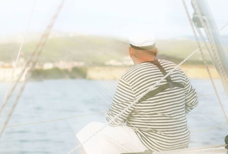 Yachtsman in marine clothing looking into the distance sitting on board a sailing yacht. Sea fishing from the ship. Peaceful state at summer holidays. Travel experienced person around the world.の写真素材