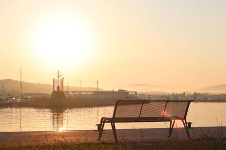 The sailing yacht enters under the motor in the marina in the rays of the morning sun. Dawn in the port. Empty bench on the pier against the lighthouse. Symbols of return and waiting. A calm romantic atmosphere.の写真素材