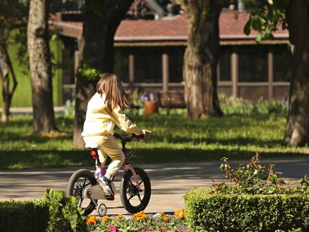 A little girl riding a bike in the park with safety wheels. Active development of the child in the fresh air. Learning to keep balance in transport management. Two-wheel balancing. Joyful childhood.の写真素材