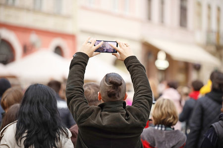 A young guy with a stylish haircut takes pictures in the crowd on his mobile phone, stretching his arms high. A fan at a concert shoots a video of his favorite band's performance on his deviceの写真素材