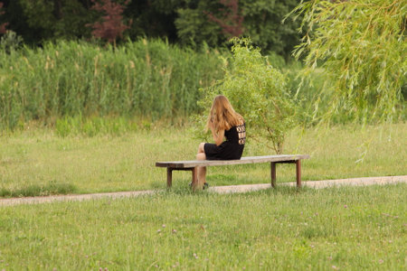 A teenage girl is sitting on a bench in a park in a black dress with a cell phone. Fashion of modern youth. Young girl relaxes in nature in a sunny park.の写真素材