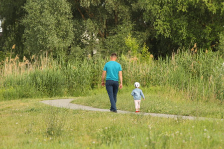 A father and his little child are walking along the alley of a summer park. A man and a girl are walking in nature. Relaxation in nature for the harmonious development of a child. Relationship betweenの写真素材