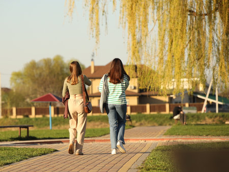 Two young girls dressed in fashionable clothes walk through a spring park in sunny weather. Girlfriends talking in nature. Walk in the fresh air.の写真素材
