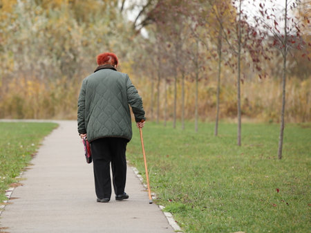 A lonely elderly woman with red hair walks with a stick along the alley of an autumn park. Breathe fresh air in nature. Active aging. Healthy lifestyle in old ageの写真素材