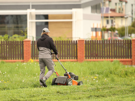 Worker using a manual lawn mower mows grass on near the house. Landscaping and maintenance of green spaces of the residential complex. Making grass lawn in a housing estate. Human labor.の写真素材