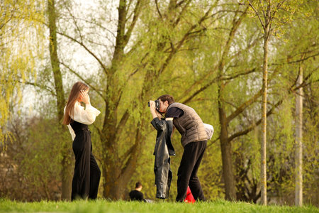Photoshoot of a young model in nature. A young photographer takes a picture of a young girl in the greenery of a summer park. The woman poses against the background of green treesの写真素材