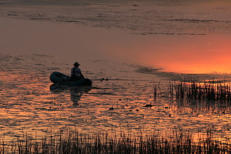 A man catches fish on an inflatable boat on the river in the evening with the setting sun. A beautiful evening in nature to relax while fishing. Calmness from the noise of city lifeの写真素材