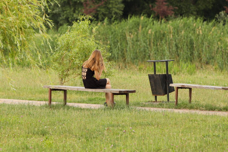 A teenage girl is sitting on a bench in a park in a black dress with a cell phone. Fashion of modern youth. Young girl relaxes in nature in a sunny park.の写真素材