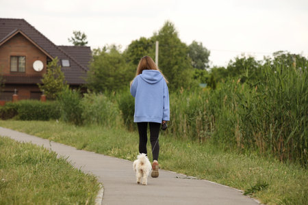 A young girl in a blue jacket and black pants walks home with her dog along an alley among the greenery. Returning from a walk with a pet. Animal care.の写真素材