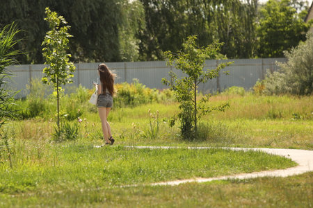 A young girl in a revealing outfit with bare legs walks along a park alley. A walk in the fresh air among summer greenery. Fashion of modern youth.の写真素材