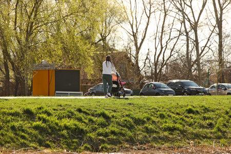 A young mother walks in the park with a baby carriage on a sunny day. Baby's stay in the fresh air. Raising the younger generation. Guardianship of human offspring.の写真素材