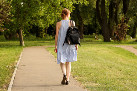 A young girl in a summer blue sundress with a leather fashionable backpack walks along a park alley among the greenery. Sophisticated fashion of modern youth.の写真素材