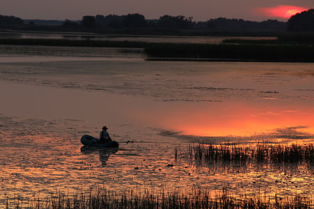 A man catches fish on an inflatable boat on the river in the evening with the setting sun. A beautiful evening in nature to relax while fishing. Calmness from the noise of city lifeの写真素材