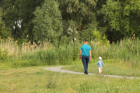 A father and his little child are walking along the alley of a summer park. A man and a girl are walking in nature. Relaxation in nature for the harmonious development of a child. Relationship betweenの写真素材