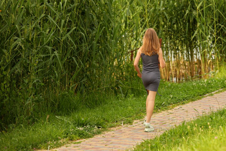 A young girl with a mobile phone in her hands walks along a park alley among the greenery to meet...