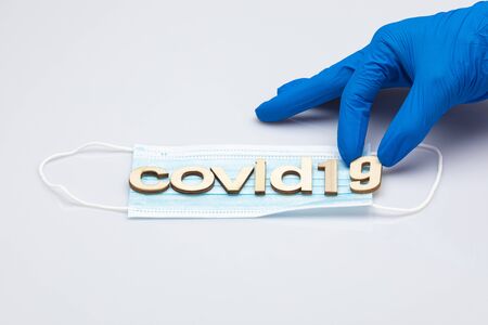 hand with a blue surgical glove holding the last part of the word covid19 formed with wooden letters placed on a surgical mask isolated on a white backgroundの写真素材