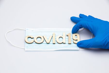 hand with a blue surgical glove holding the last part of the word covid19 formed with wooden letters placed on a surgical mask isolated on a white backgroundの写真素材