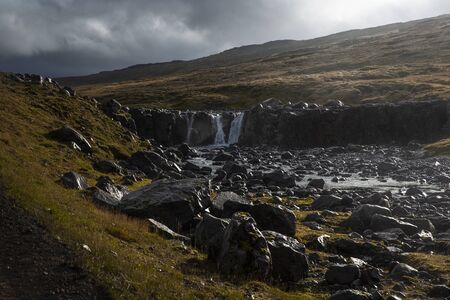 landscape with a visible waterfall in Iceland. The water is clear blue and is surrounded by dark volcanic stones full of mossの写真素材