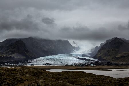 Mountain landscape in Iceland. in the distance you can see the Sólheimajökull glacier with a sky very cloudy above. The sky is not visible and no ray of sunshine passes throughの写真素材