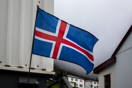 landscape in the town of Ólafsfjörður, in the north of iceland. An Icelandic flag attached to the edge of the window of a house flies in the wind. The sky is loaded with cloudsの写真素材