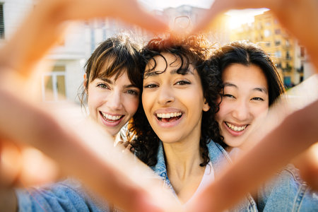 Three happy diverse young women drawing heart shape with hands outdoorsの写真素材