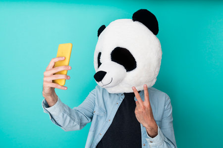 Happy man in panda mask taking a selfie with smartphone while gesturing victory sign over blue backgroundの写真素材