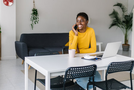 Happy young african woman using mobile phone while working on laptop at homeの写真素材