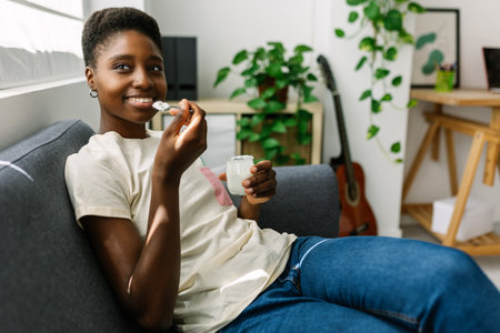 Happy young african woman relaxing on sofa eating organic yogurtの写真素材