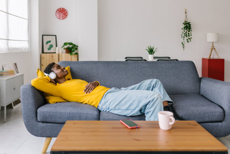 Happy young african woman listening to music while lying on sofa at homeの写真素材