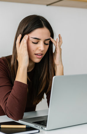 Young adult woman suffering headache or migraine while working on laptop at home