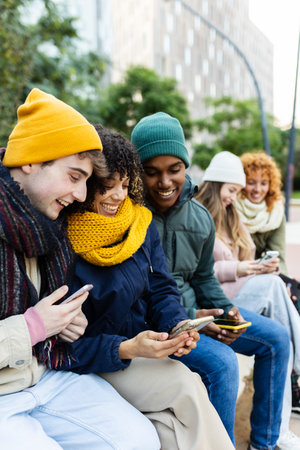 Diverse teenagers using mobile phone sitting on city bench on winter