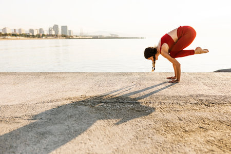 Young hispanic woman practicing yoga and doing bakasana position by the sea