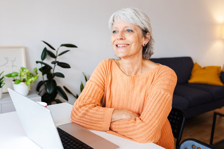 Senior woman with laptop looking away sitting on table at homeの写真素材