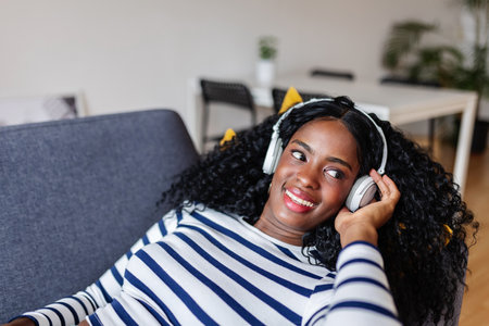 Young happy black woman enjoying online music while resting on sofa at homeの写真素材