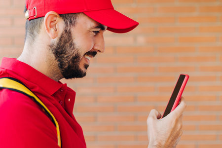Happy hispanic delivery man in red clothes using mobile phoneの写真素材