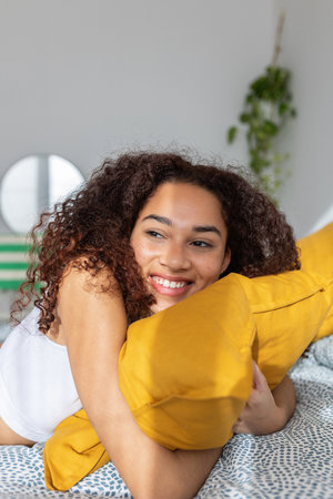 Smiling young adult woman hugging yellow pillow relaxing on bed at homeの写真素材