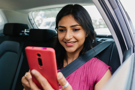 Happy young adult indian woman using mobile phone while traveling by taxi