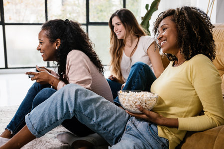Happy female friends playing videogames and eating popcorn at homeの写真素材